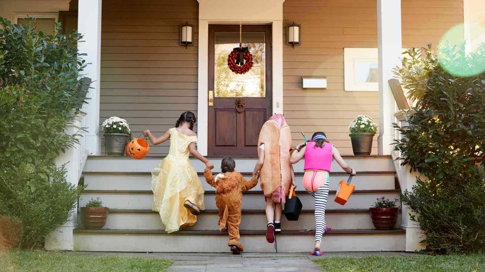 Four children in costumes walk up the steps of a porch, carrying treat buckets, likely trick-or-treating for Halloween.
