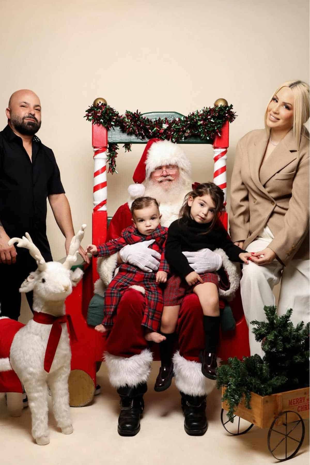 Two children sit on Santa’s lap in front of a festive sleigh, with a man, a woman, and a decorative reindeer nearby. Holiday decorations are visible.