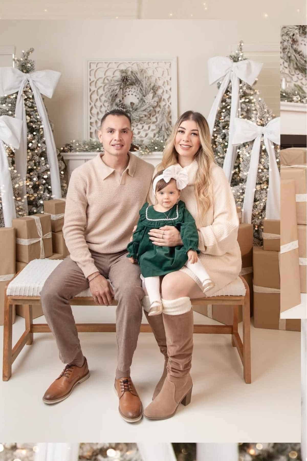 A man, woman, and young girl sit on a bench in front of decorated Christmas trees and wrapped gifts, all dressed in neutral and green festive attire.