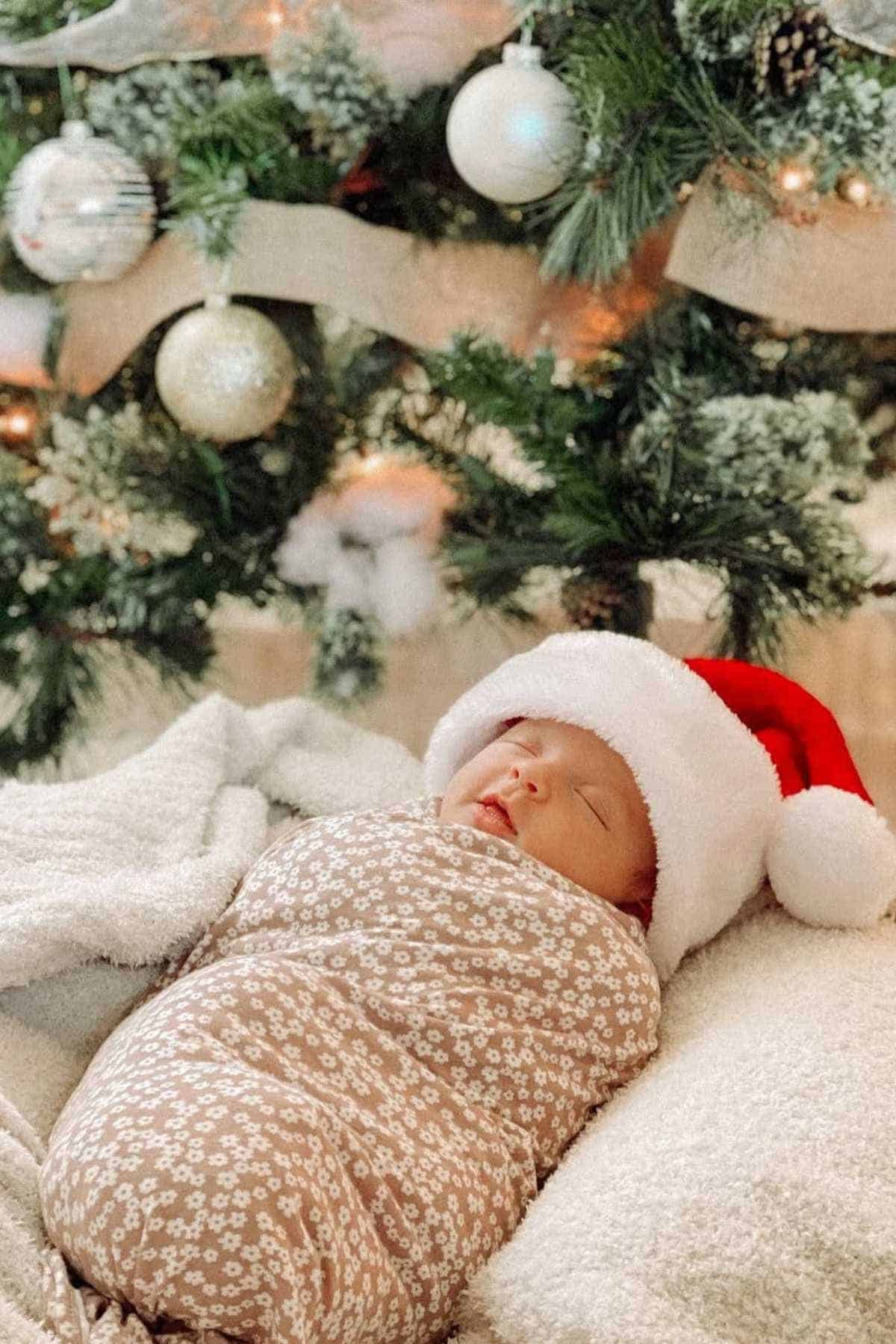 A newborn baby wrapped in a floral blanket sleeps under a Christmas tree, wearing a Santa hat.