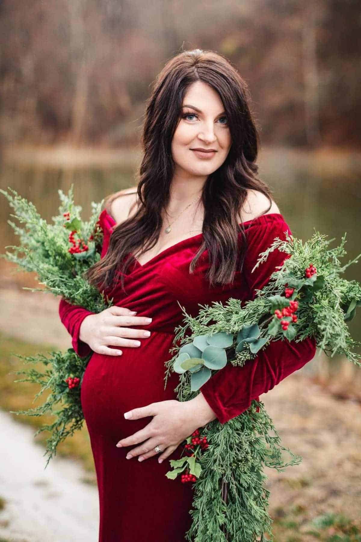 Pregnant woman in a red velvet dress stands outdoors, holding her belly and decorated with green foliage and red berries, with a blurred natural background.