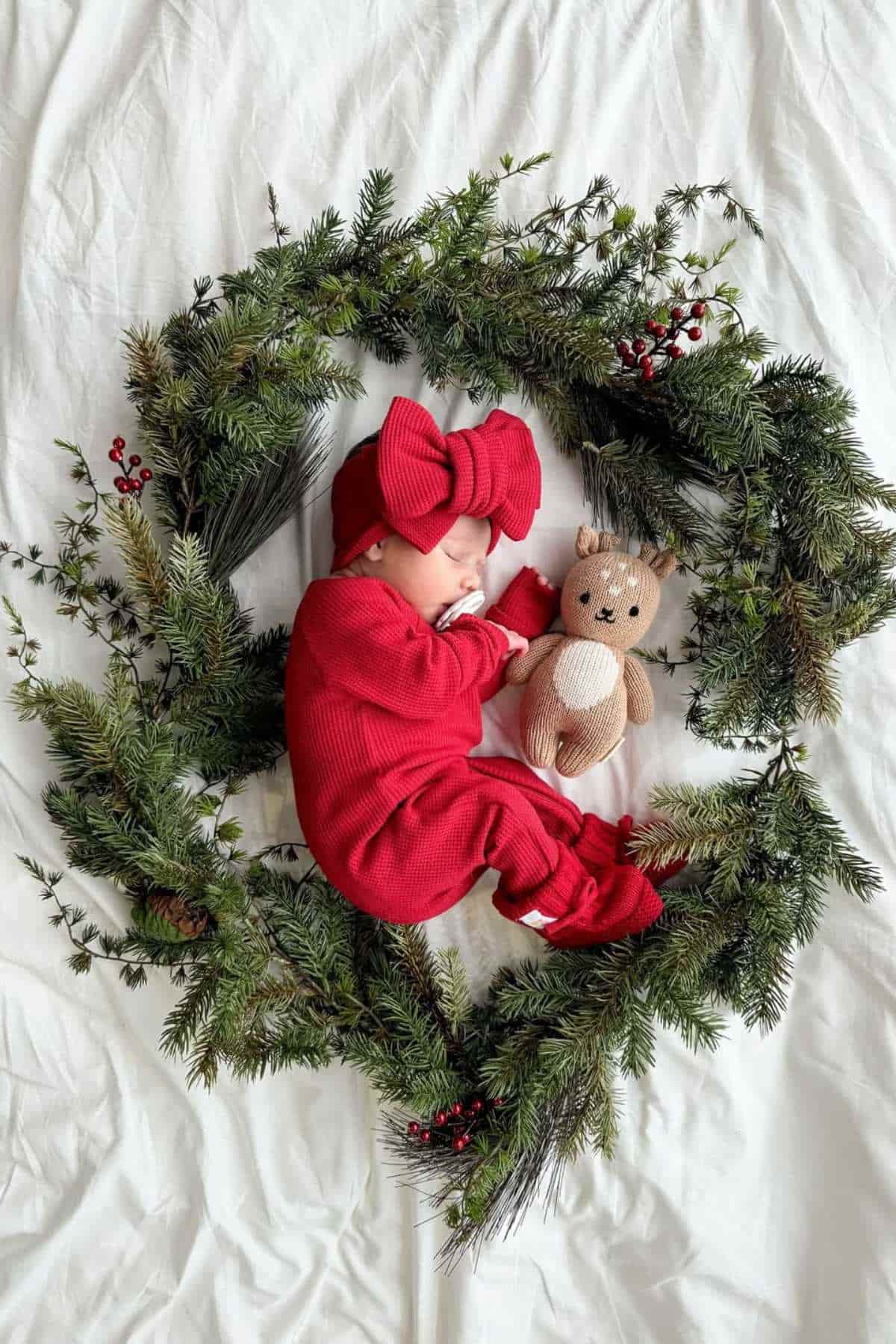 A baby dressed in red with a large bow sleeps on a white blanket, holding a stuffed bear, surrounded by a circular wreath of greenery and berries.
