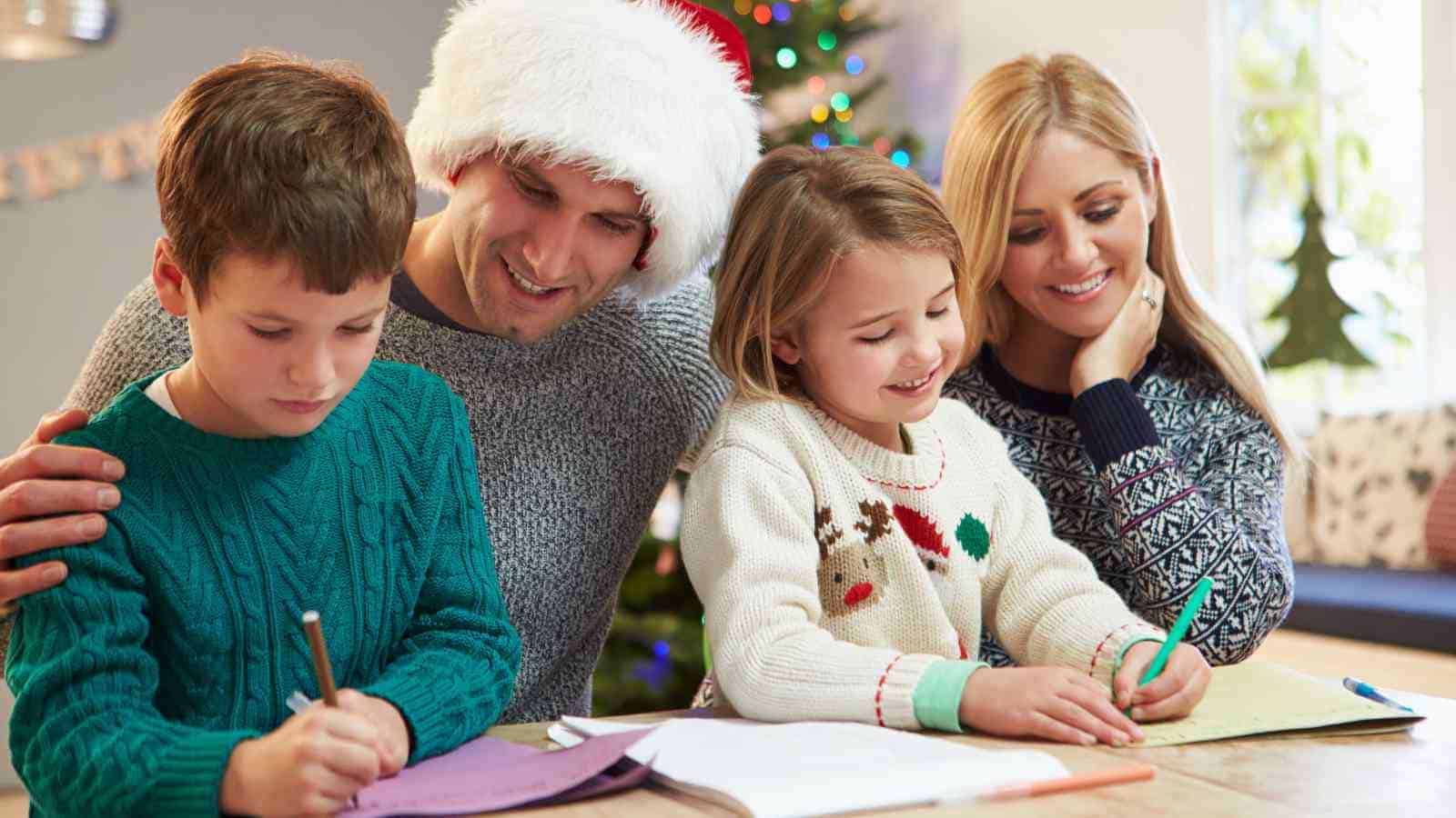 A family of four sits at a table doing holiday crafts, with a Christmas tree in the background. The father wears a Santa hat.