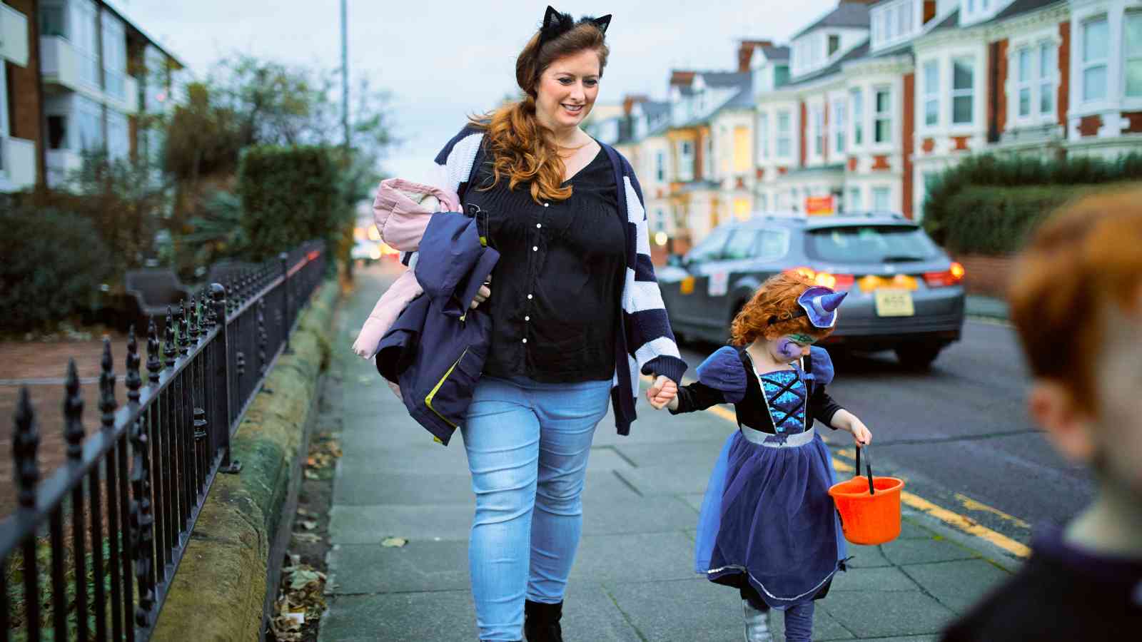 An adult and a child in costumes walk on a sidewalk; the child carries an orange bucket. Cars and houses line the street in the background.