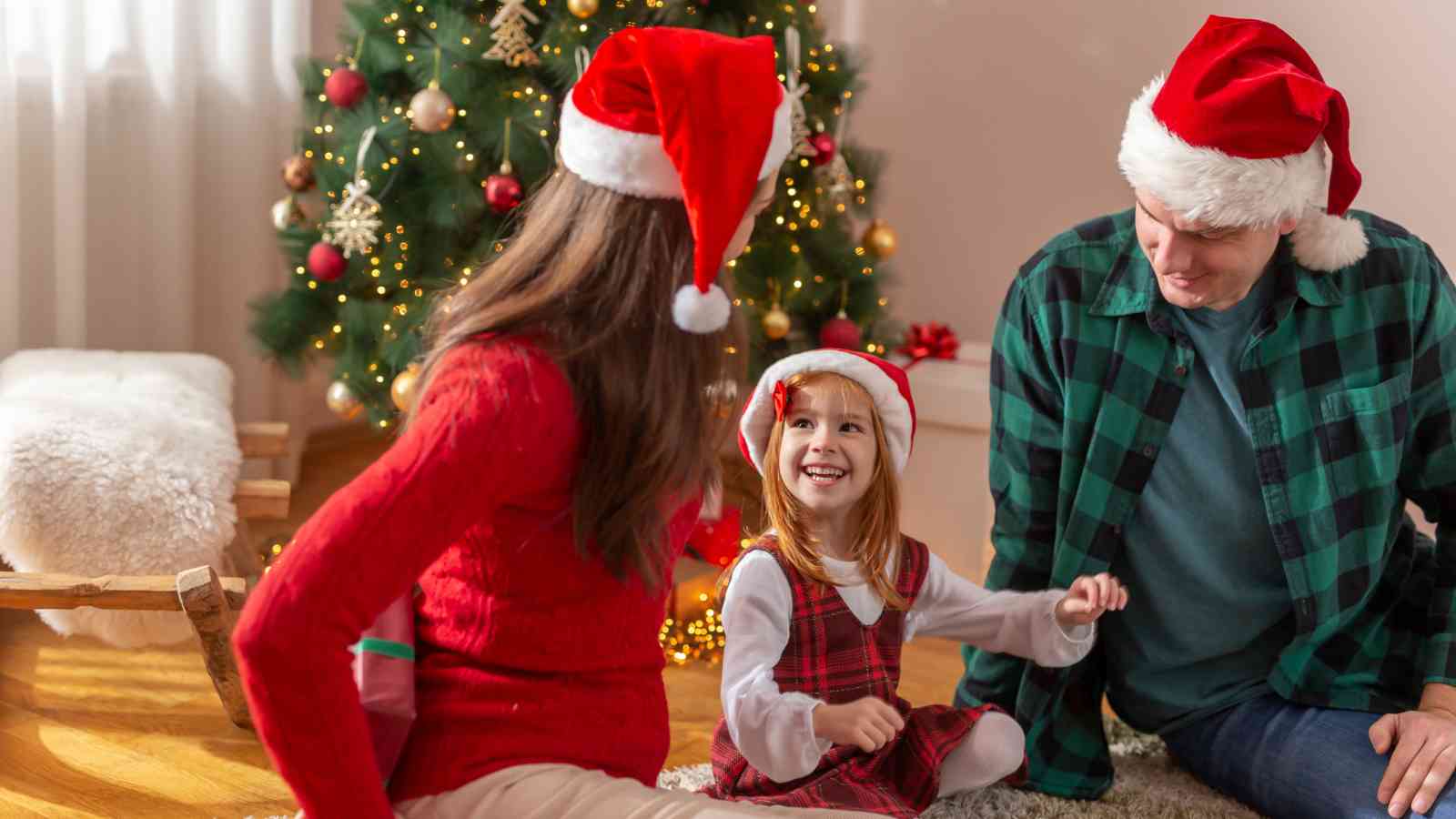 A family wearing Santa hats sits on the floor near a decorated Christmas tree, with a young girl smiling at the adults.