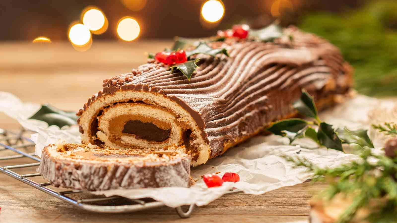A chocolate Yule log cake with decorative frosting, holly leaves, and berries, is partially sliced and rests on parchment paper atop a cooling rack.