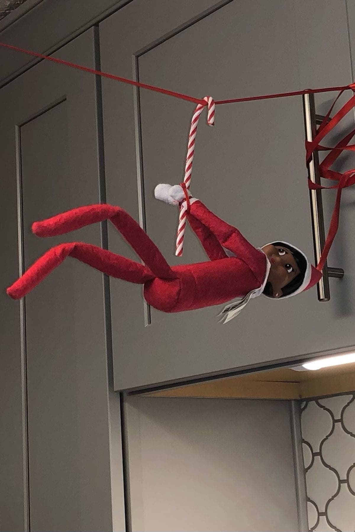 A toy elf doll in red clothes is hanging from a red ribbon zipline, holding a candy cane, against a kitchen cabinet background.