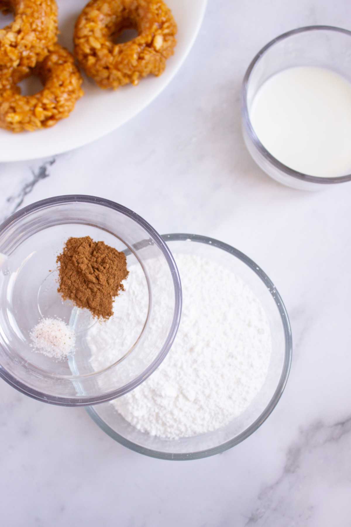 Three glass bowls filled with powdered sugar, spices, and milk are arranged on a marble surface; a plate of pumpkin rice krispies donuts is partially visible in the corner.
