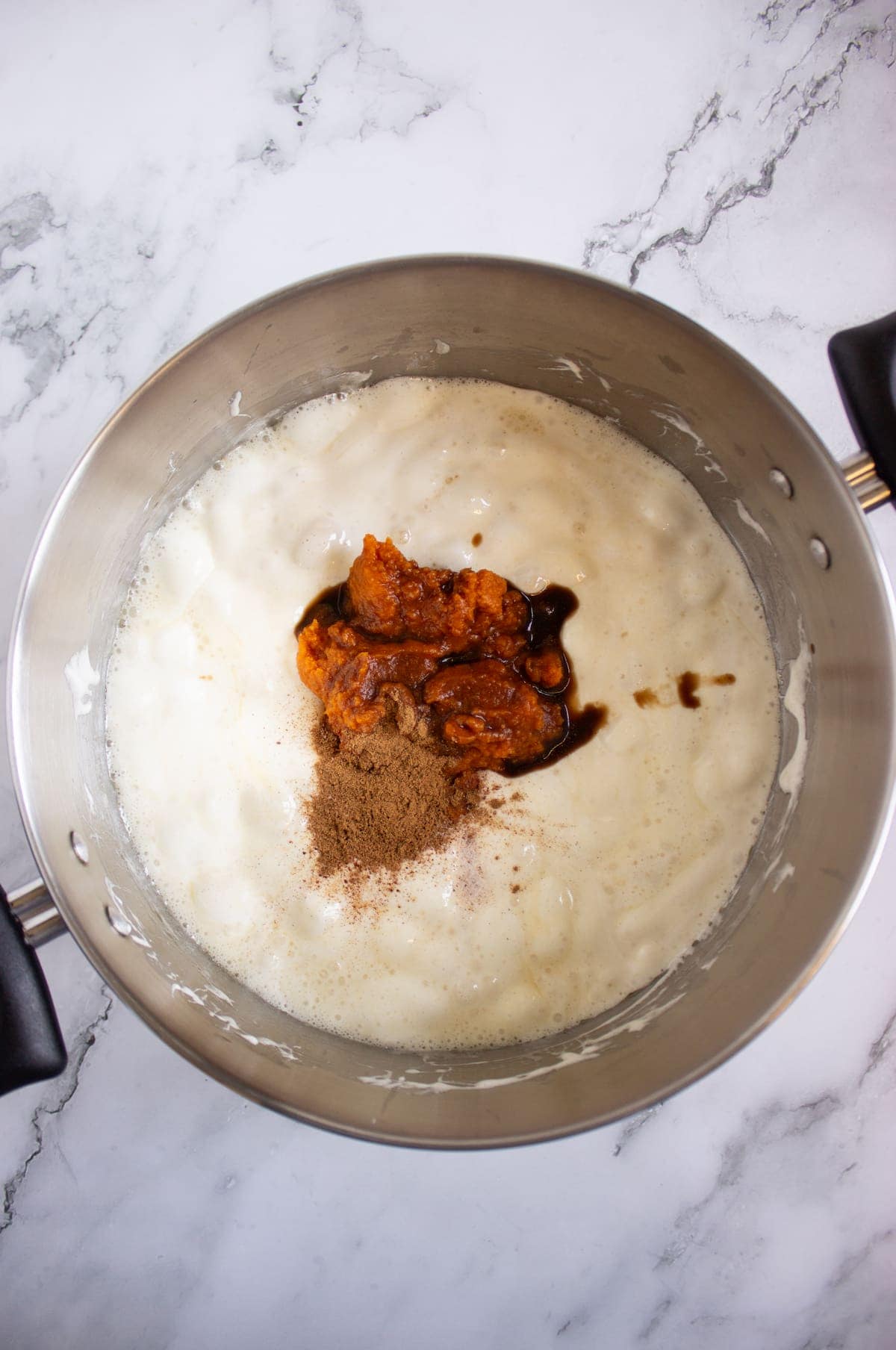 A metal pot containing a creamy mixture with dollops of pumpkin puree, brown spices, and dark liquid on a marble countertop.