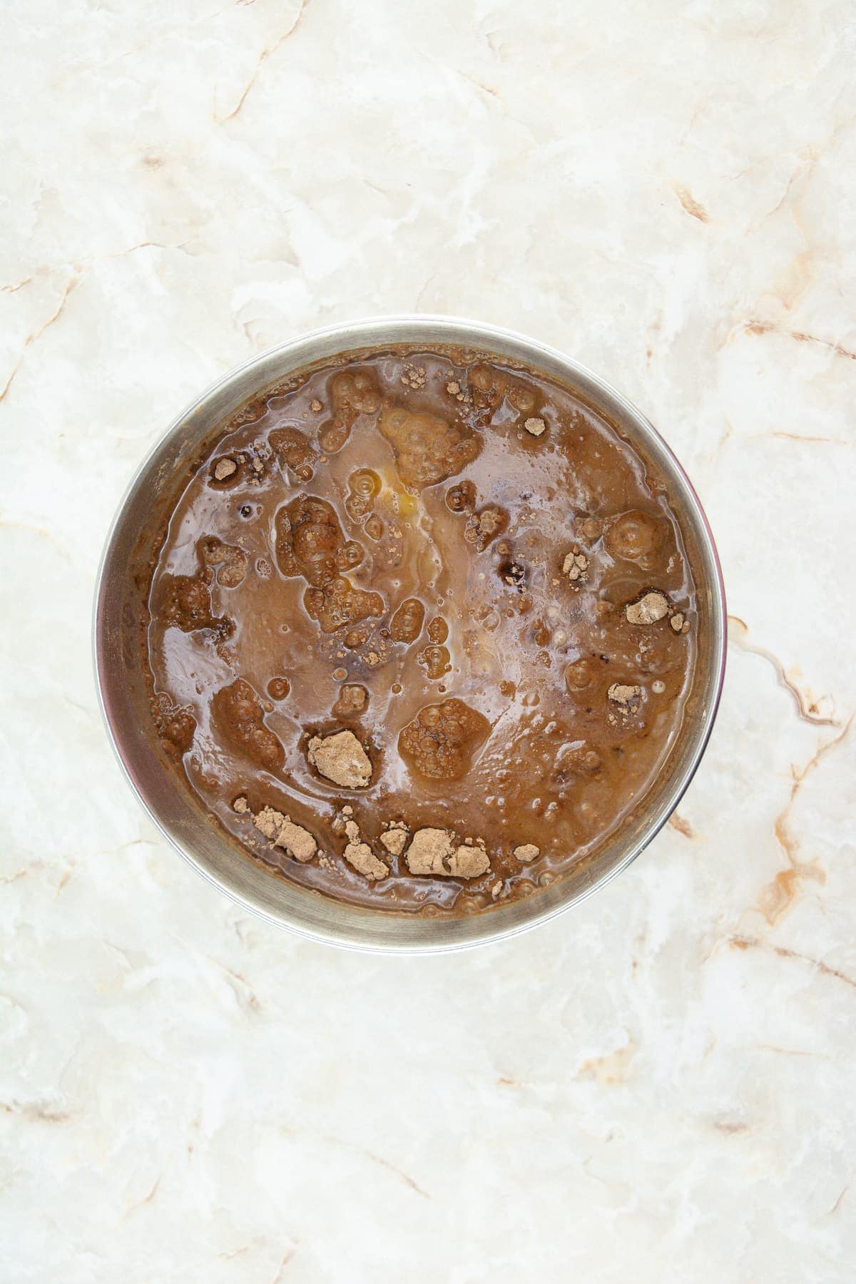 A glass bowl containing a mixture of cocoa powder, sugar, and liquid on a light marble surface.