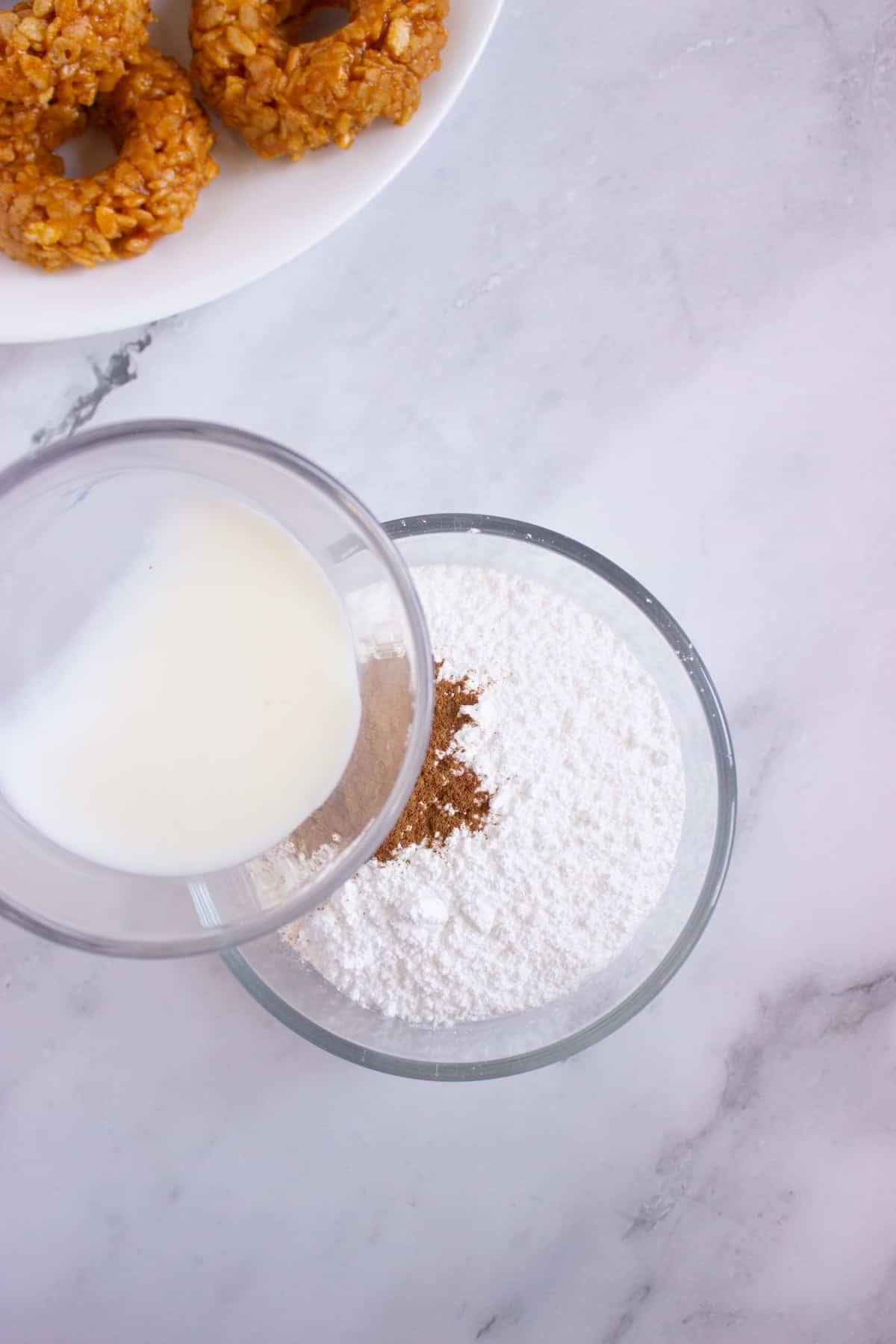 A glass of milk being poured into a bowl containing powdered sugar and spices, with two donuts on a white plate in the corner.