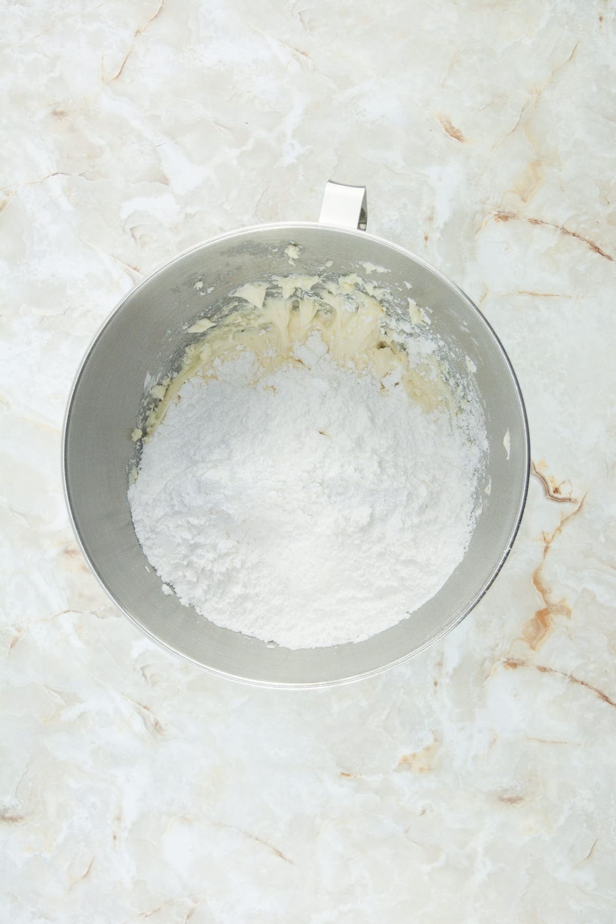 A metal mixing bowl containing powdered sugar and butter on a light marble surface.