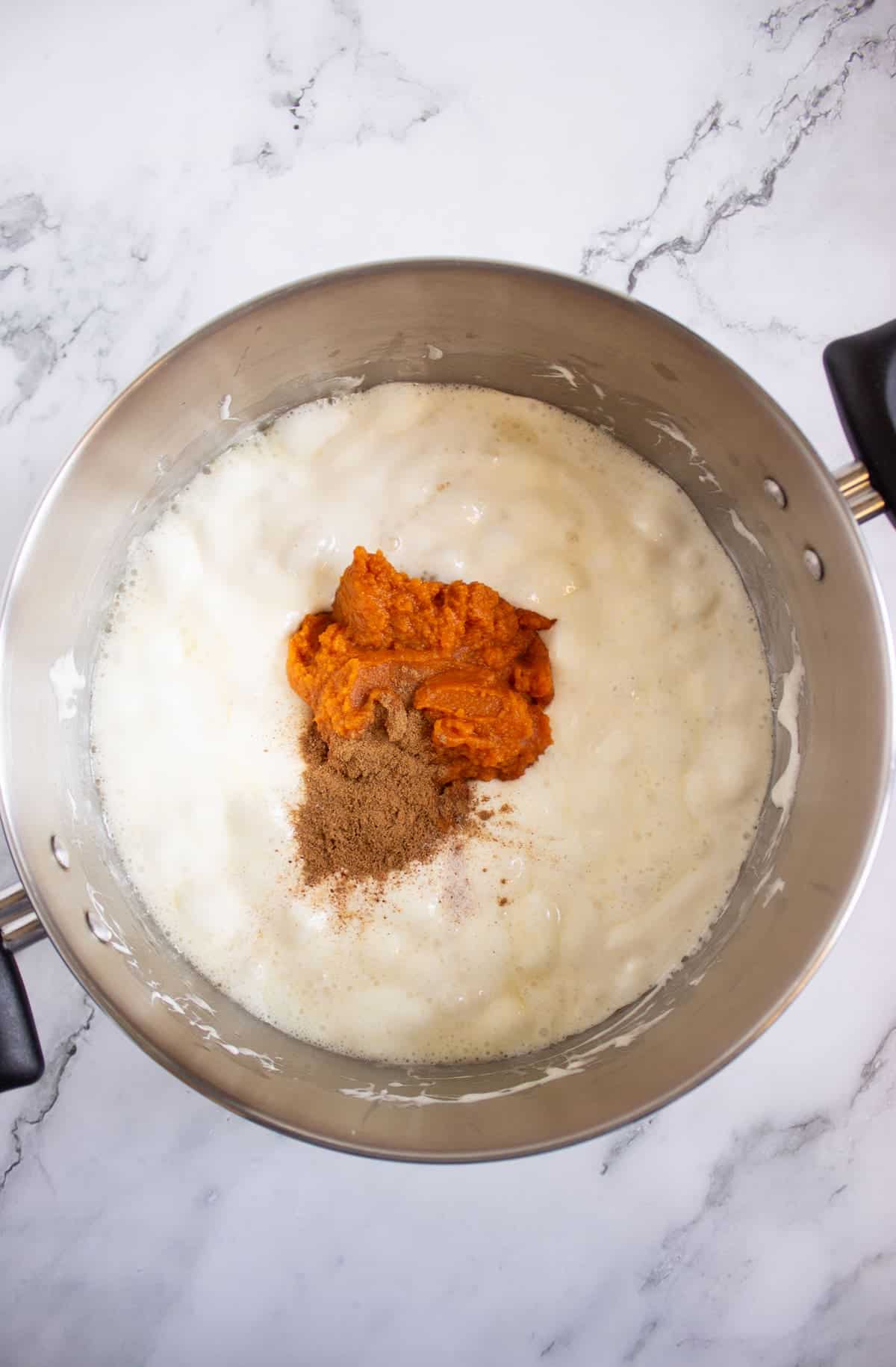 A saucepan filled with a creamy mixture, topped with a dollop of pumpkin puree and a sprinkle of spices, on a marble countertop.