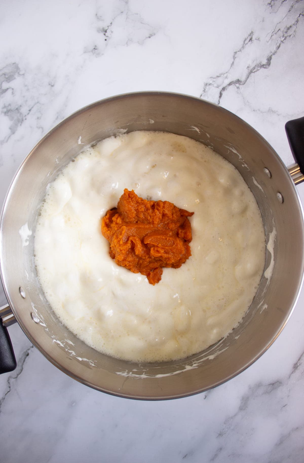 A stainless steel pot containing a white creamy mixture with a dollop of orange puree in the center, on a marble countertop.