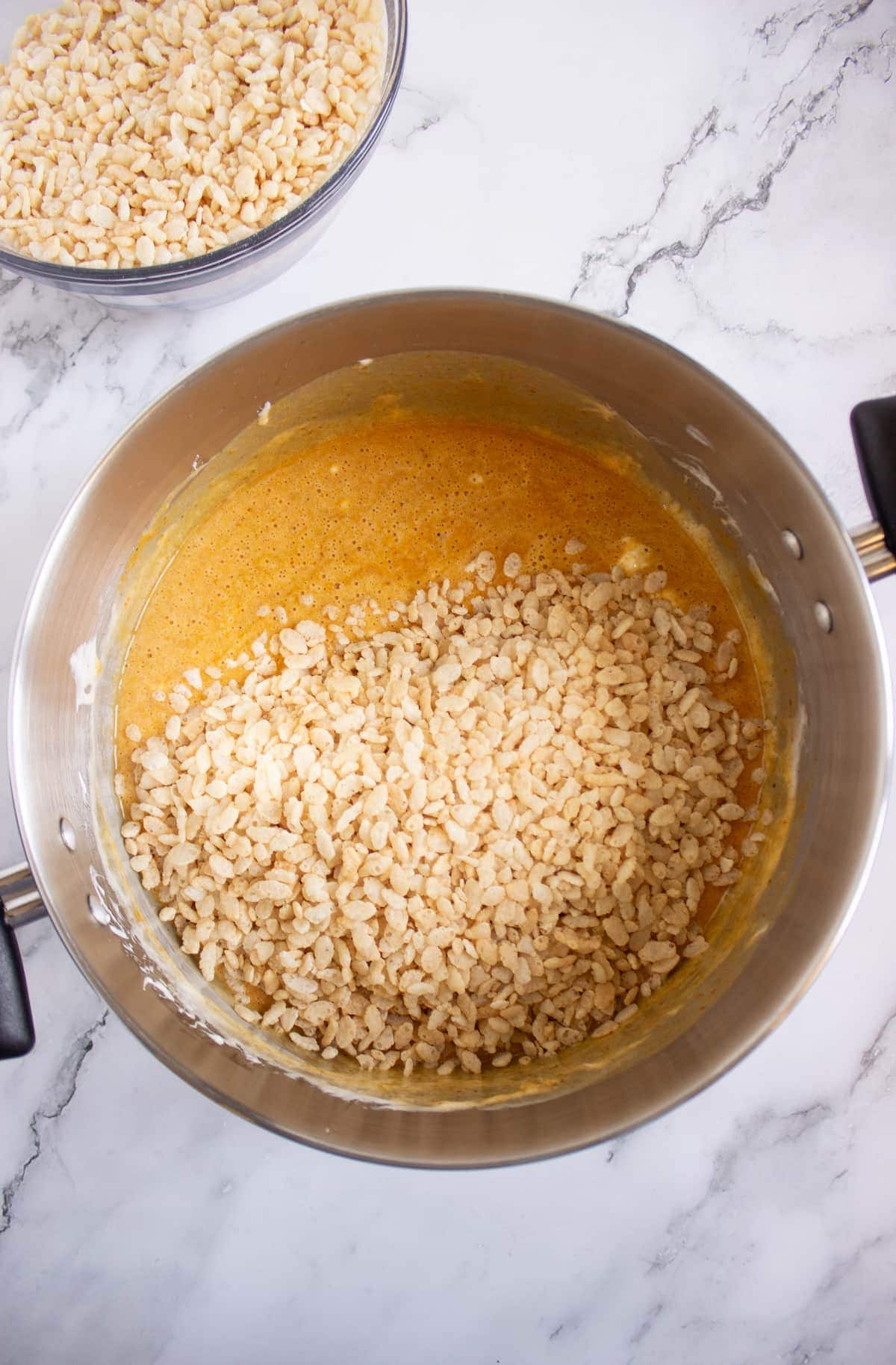 A large pot with melted marshmallow mixture and rice cereal being combined, with an additional bowl of rice cereal in the background.