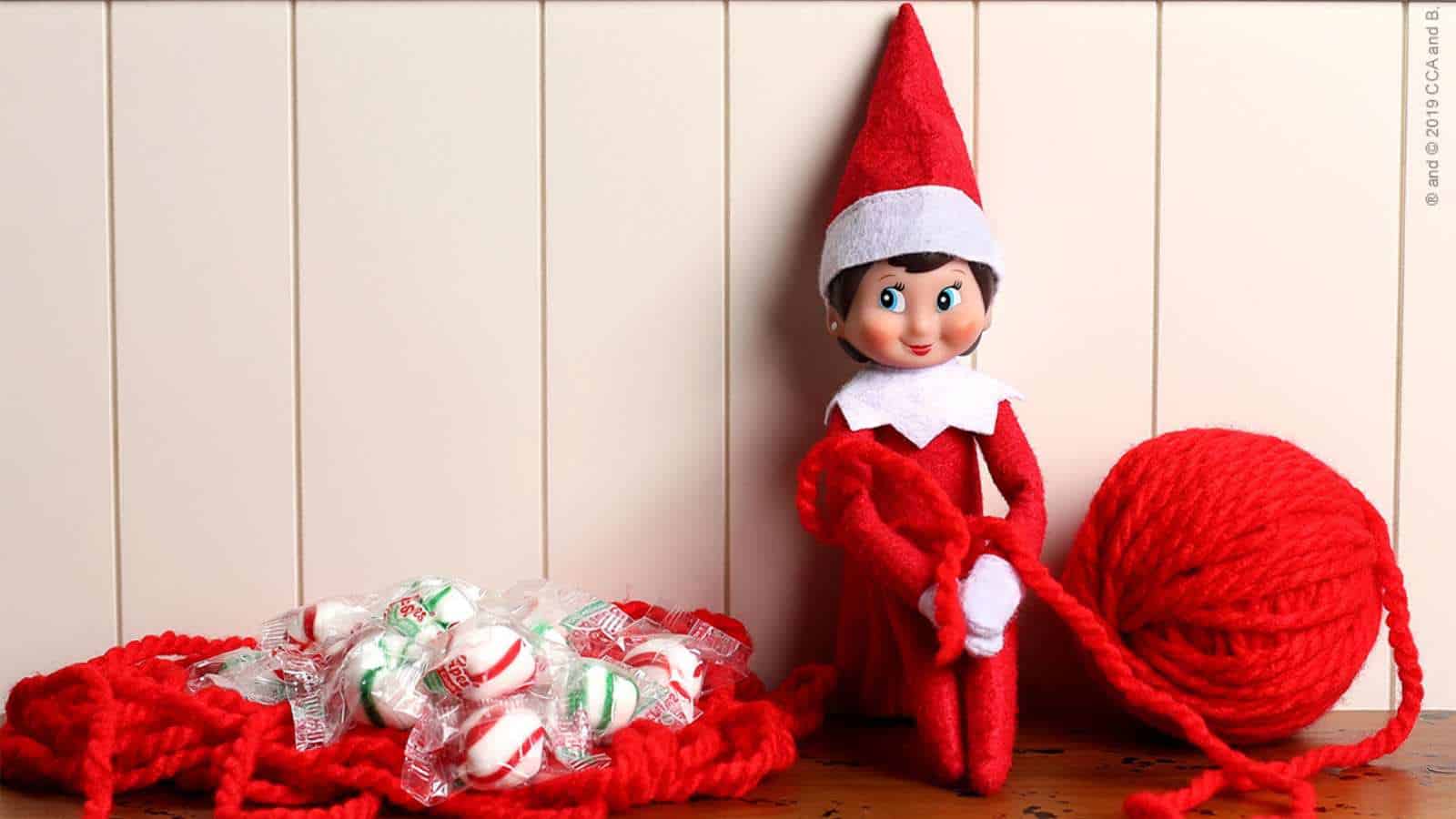A toy elf in a red outfit sits next to a ball of red yarn, surrounded by red yarn loops and individually wrapped peppermint candies.