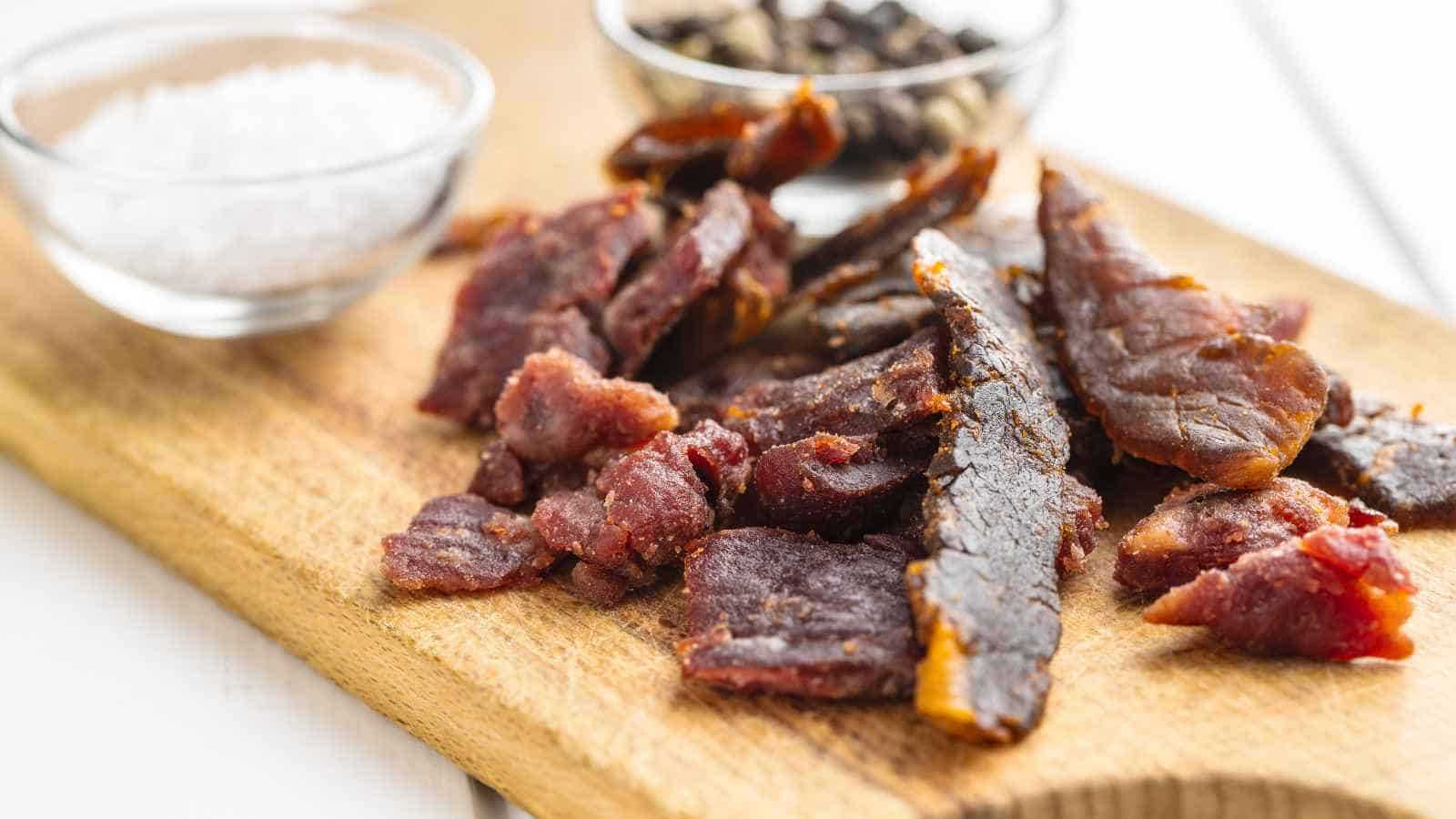 Sliced beef jerky on a wooden cutting board with small glass bowls of coarse salt and whole black pepper in the background.