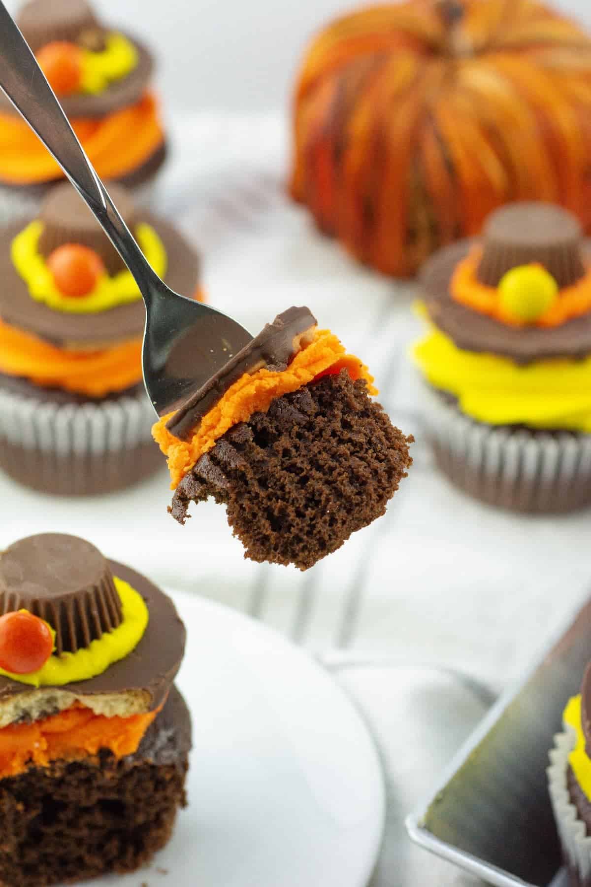 A fork holds a bite of chocolate cupcake with orange frosting. In the background, decorated cupcakes and a pumpkin are visible.