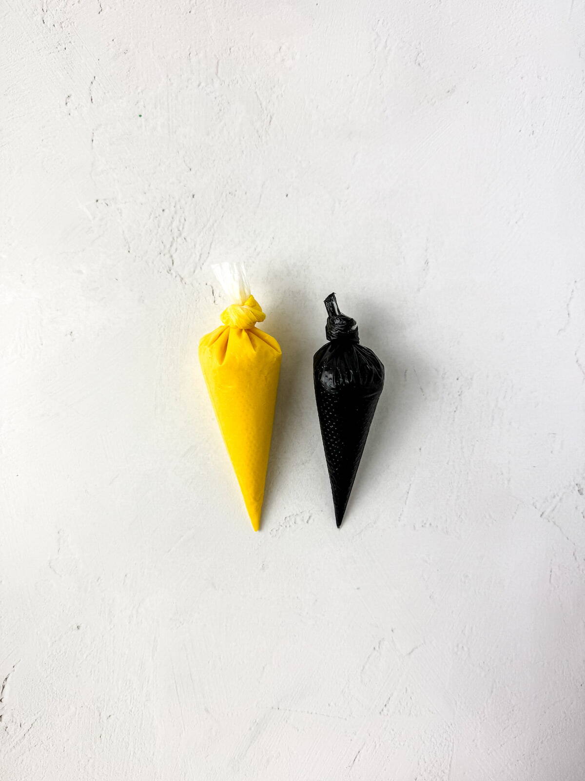 Two small piping bags, one filled with yellow icing and the other with black icing, placed side by side on a white textured surface.