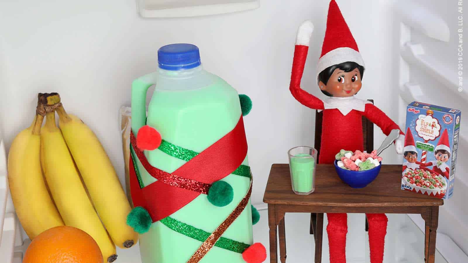 A toy elf sits at a small table with a bowl of cereal and green milk inside a refrigerator, next to bananas, an orange, and a decorated milk jug.