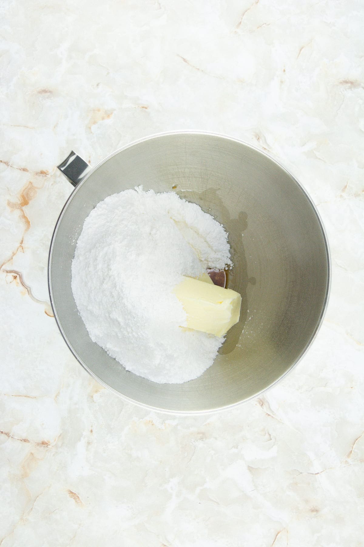A mixing bowl containing powdered sugar, a stick of butter, and a small amount of liquid on a marble countertop.