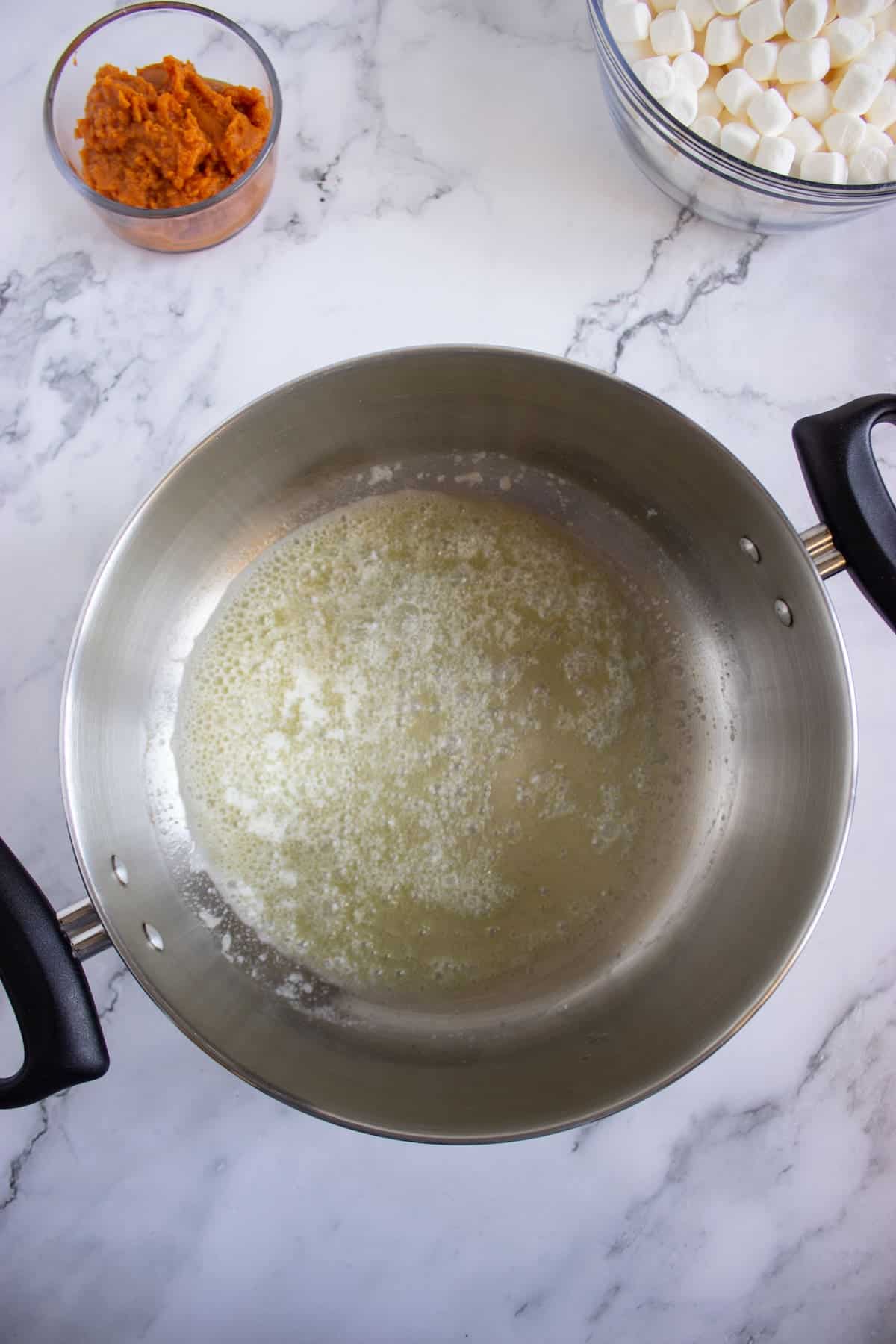 A pot with melted butter sits on a marble countertop, with bowls of mini marshmallows and orange sweet potato puree nearby.