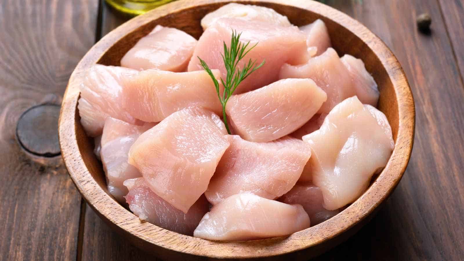 A wooden bowl filled with raw, diced chicken breast pieces, garnished with a small sprig of dill, placed on a wooden surface.