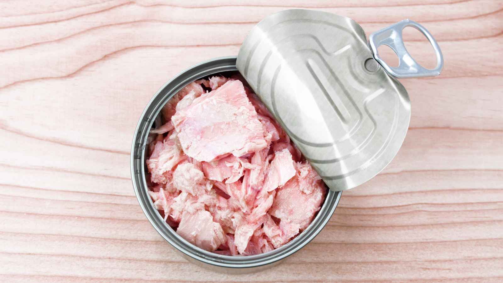 An open tin can of pink canned tuna sits on a light wooden surface, with the lid partially peeled back.