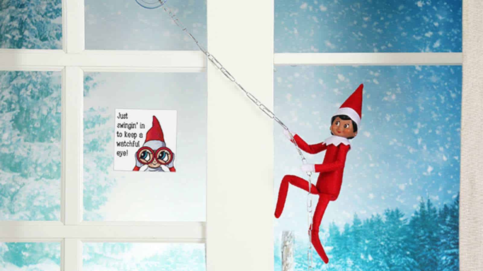 A toy elf dressed in red climbs a white window frame with a chain. Snowy trees are visible outside, and a sign on the window says, “Just swingin’ in to keep a watchful eye!”.