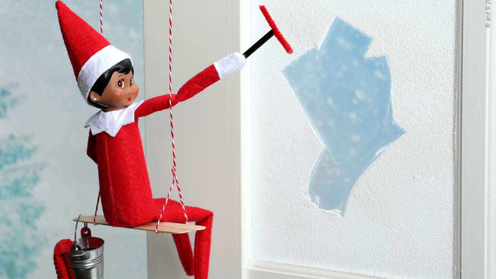 A toy elf in a red outfit sits on a swing, cleaning a window with a squeegee and bucket, leaving a clear patch on the frosted glass.