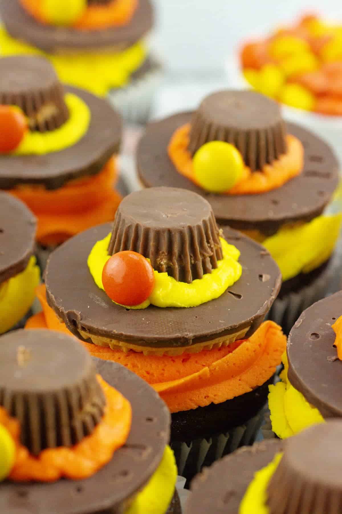 Chocolate cupcakes decorated with orange and yellow frosting, topped with a chocolate cookie, a peanut butter cup, and small round candies, resembling a Thanksgiving hat.