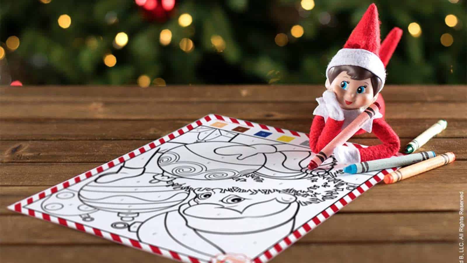 A toy elf sits on a wooden table holding a crayon next to a holiday-themed coloring sheet and several crayons, with a decorated Christmas tree in the background.