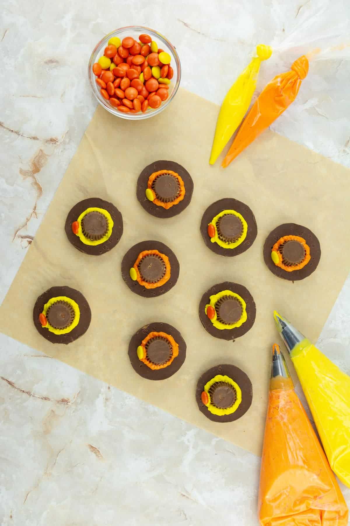 Chocolate cookies topped with peanut butter cups and orange and yellow icing rings, with piping bags and a small bowl of orange candy-coated chocolates on a marble surface.