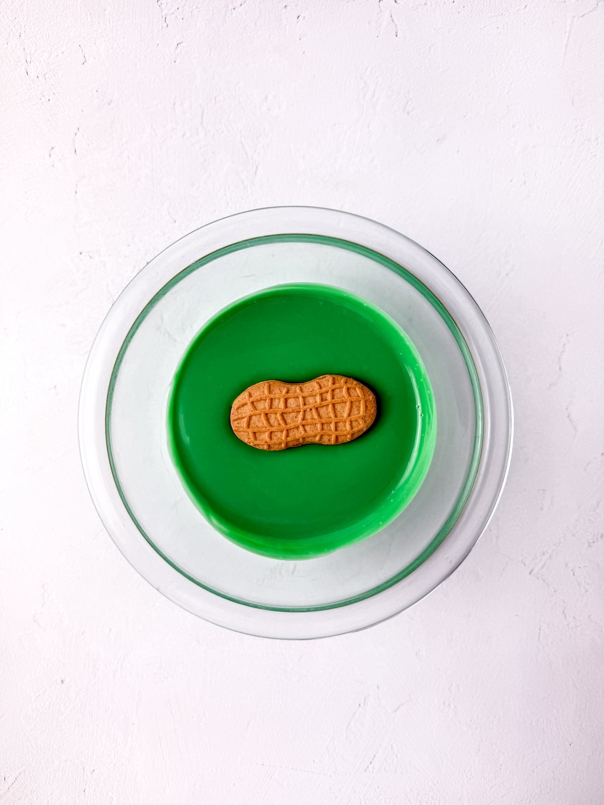 A peanut-shaped cookie sits in the center of a glass plate with green liquid, against a white textured background.