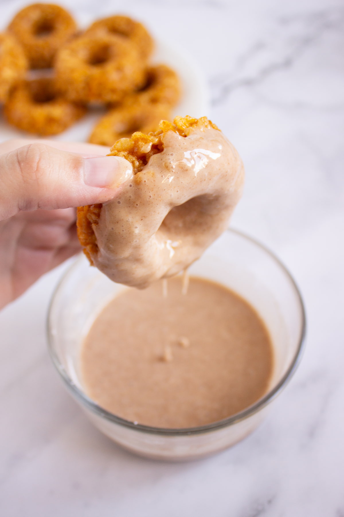 A hand dips a baked donut into a bowl of light brown glaze, with more donuts on a plate in the background.