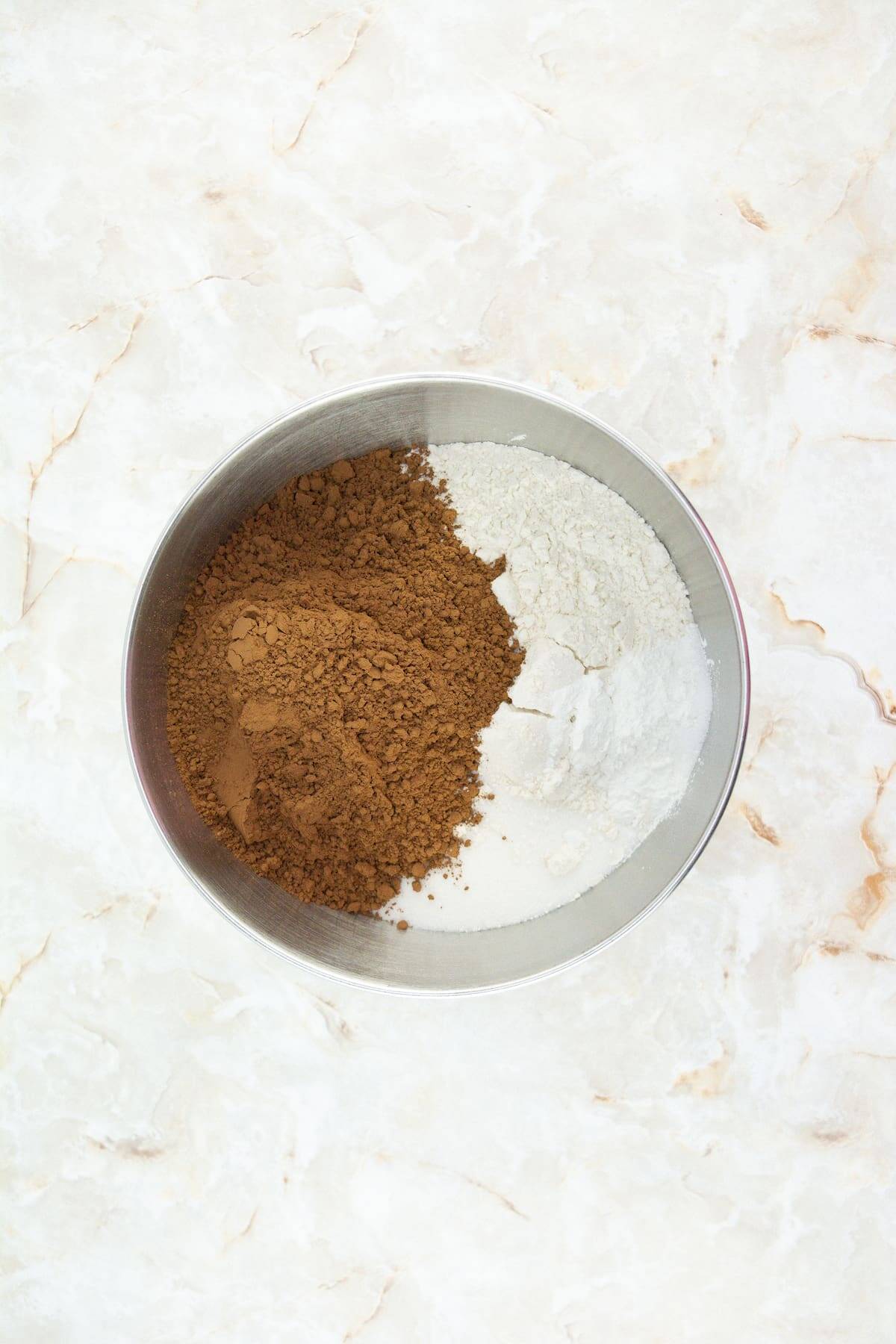 A metal bowl containing cocoa powder, flour, and granulated sugar on a light marble surface.
