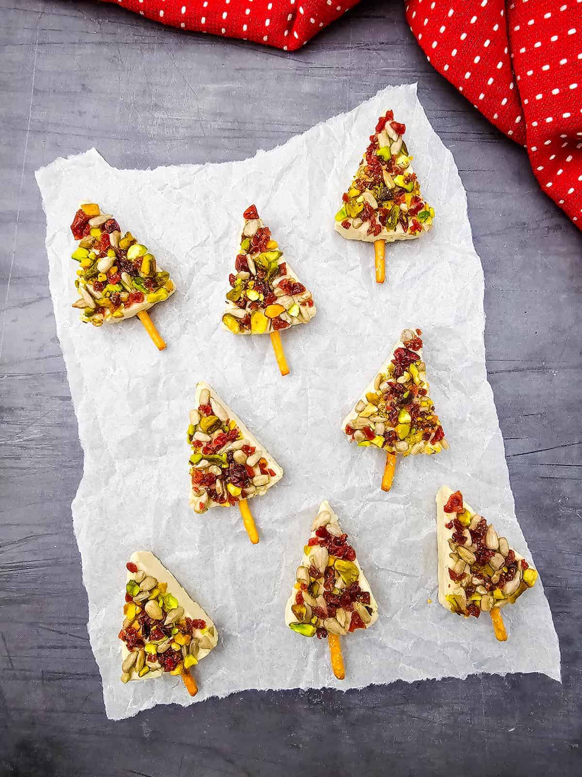 Eight Christmas tree-shaped white chocolate bark pieces with pretzel stick trunks, topped with nuts and dried fruits, arranged on parchment paper over a dark surface with a red cloth nearby.