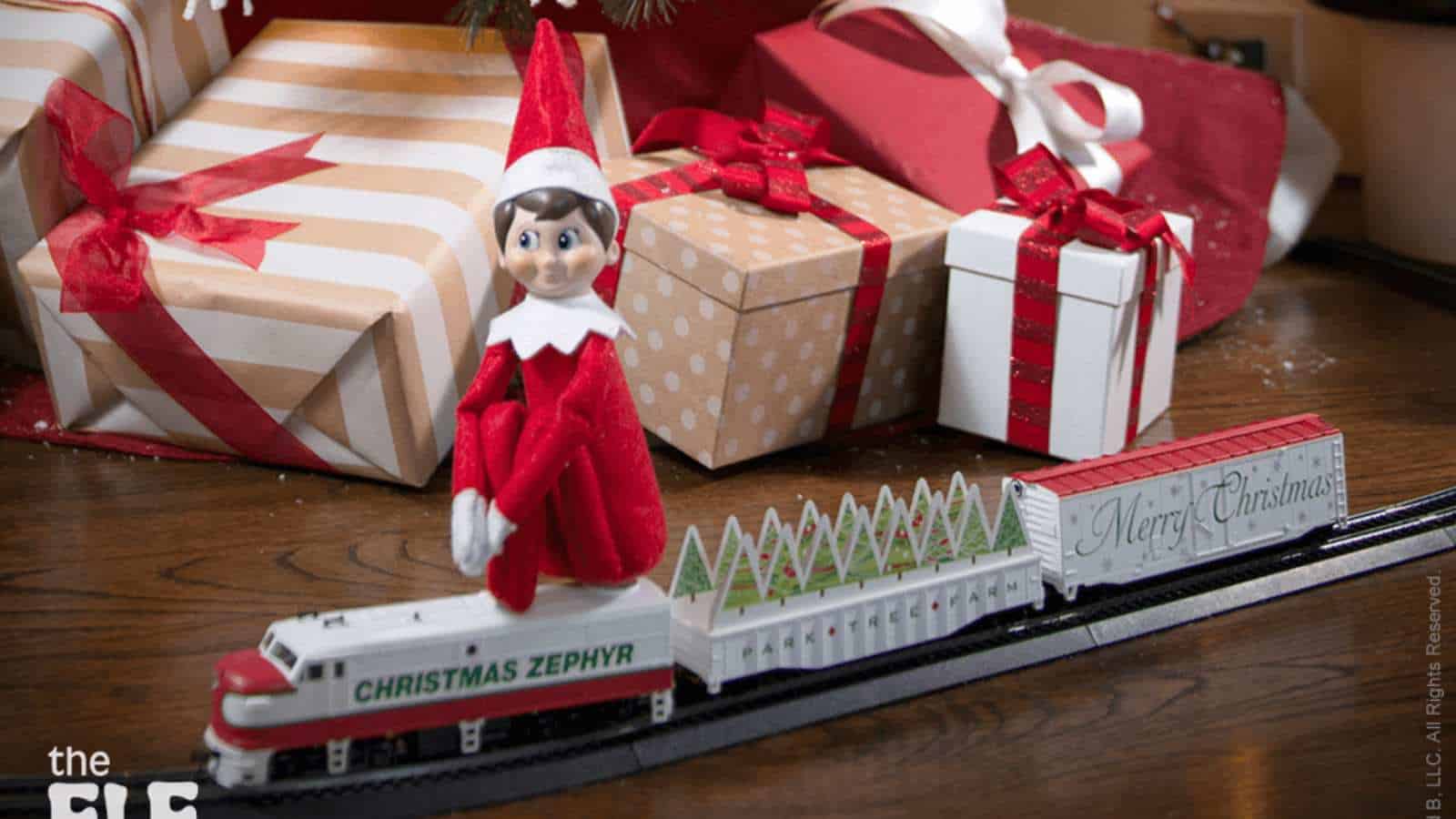 A toy elf in a red and white outfit sits on a model train labeled "Christmas Zephyr," with wrapped gifts and a Christmas tree in the background.