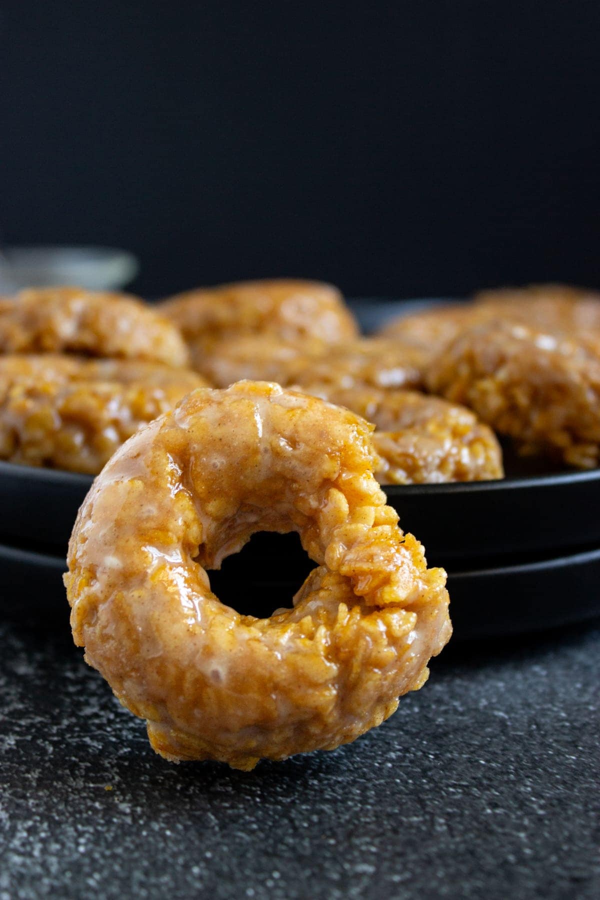 A close-up of a glazed donut with a textured surface, positioned in front of a plate stacked with more donuts against a dark background.