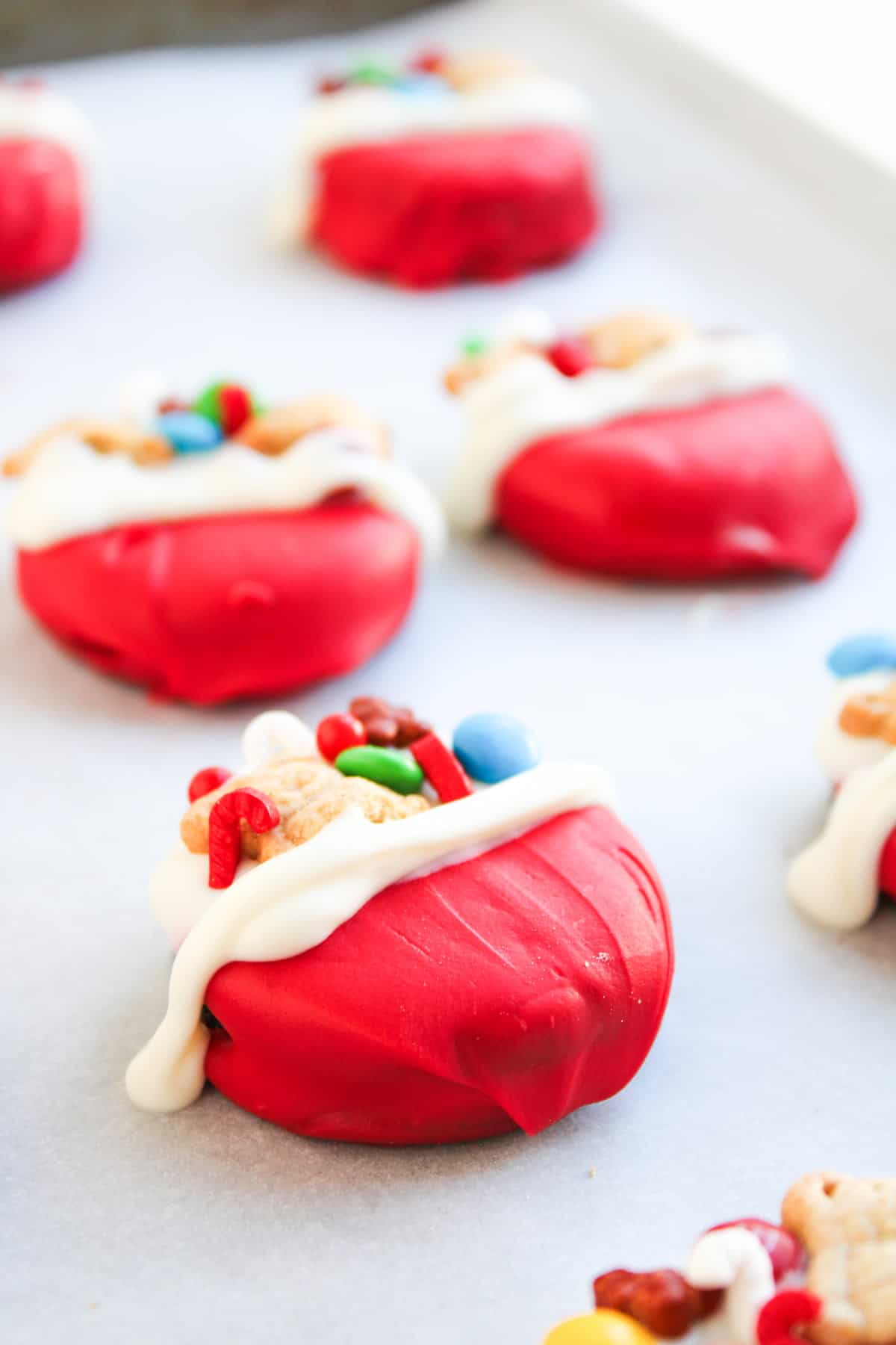 Red, round cookies decorated with white icing, mini candy pieces, and cereal, arranged on a baking sheet lined with parchment paper.