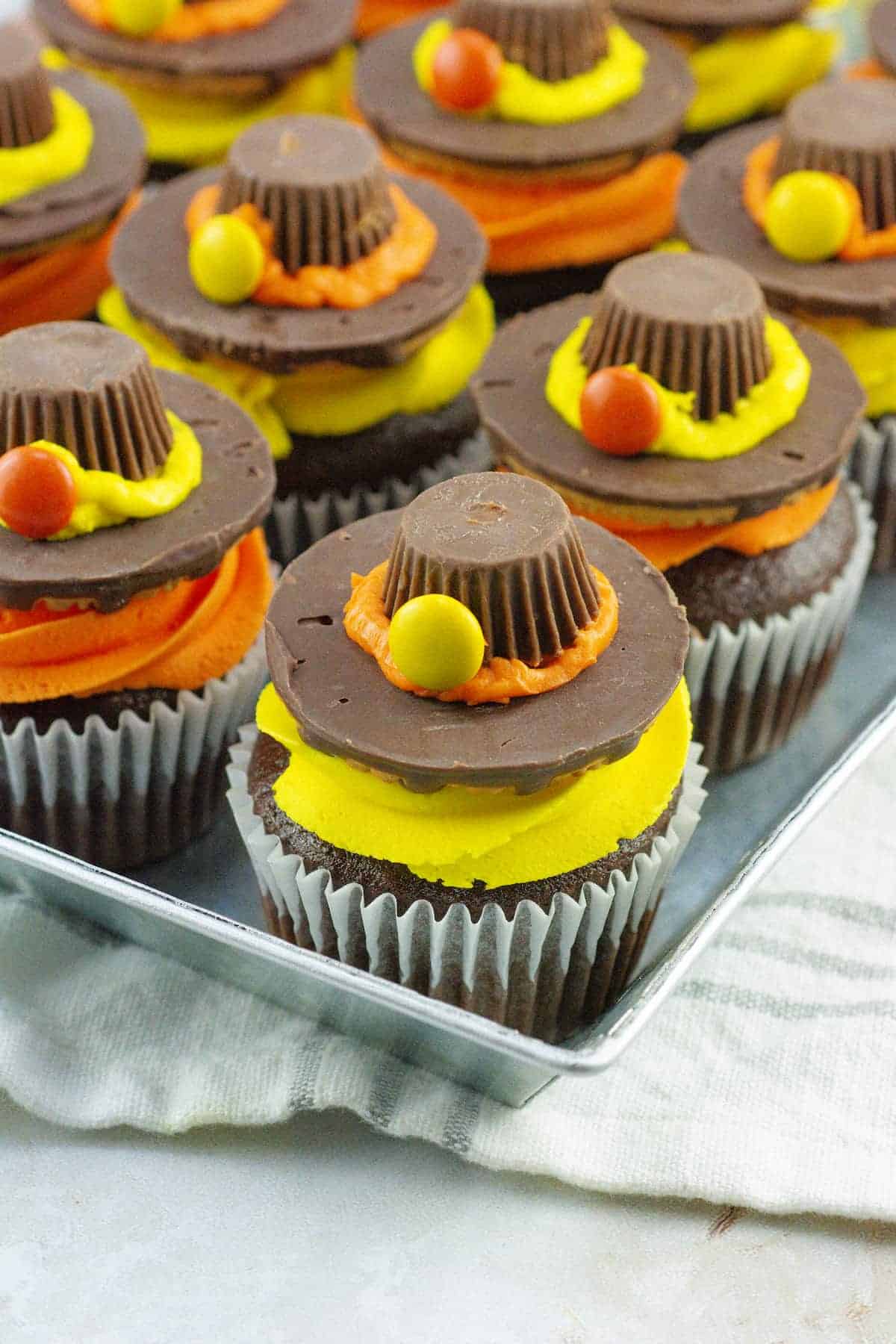 Chocolate cupcakes decorated with yellow and orange frosting, topped with a chocolate cookie, a peanut butter cup, and candy to resemble Thanksgiving hats, arranged on a metal tray.