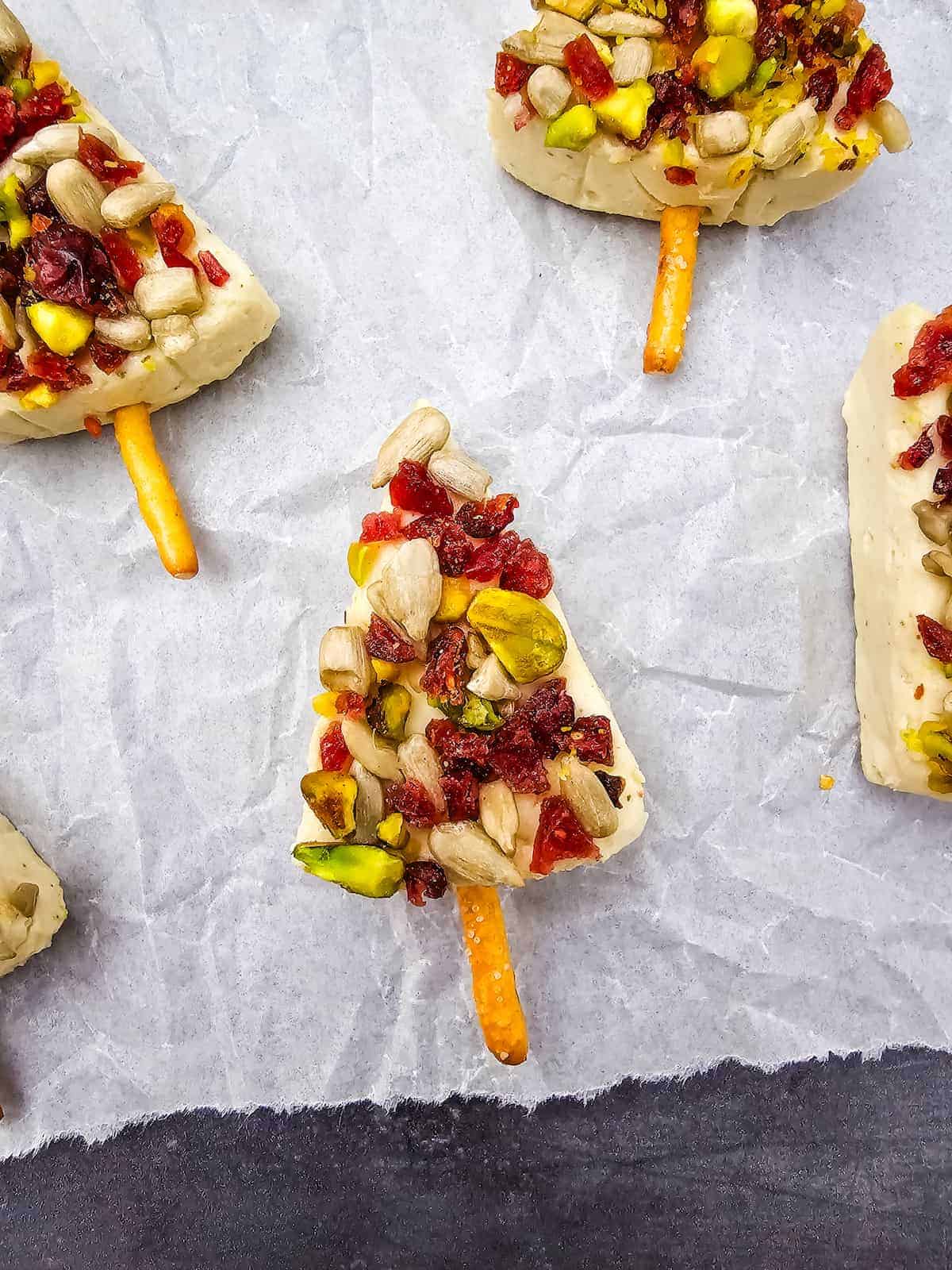 Tree-shaped cheese appetizer topped with dried cranberries, sunflower seeds, and pistachios, with a pretzel stick as the trunk, arranged on parchment paper.