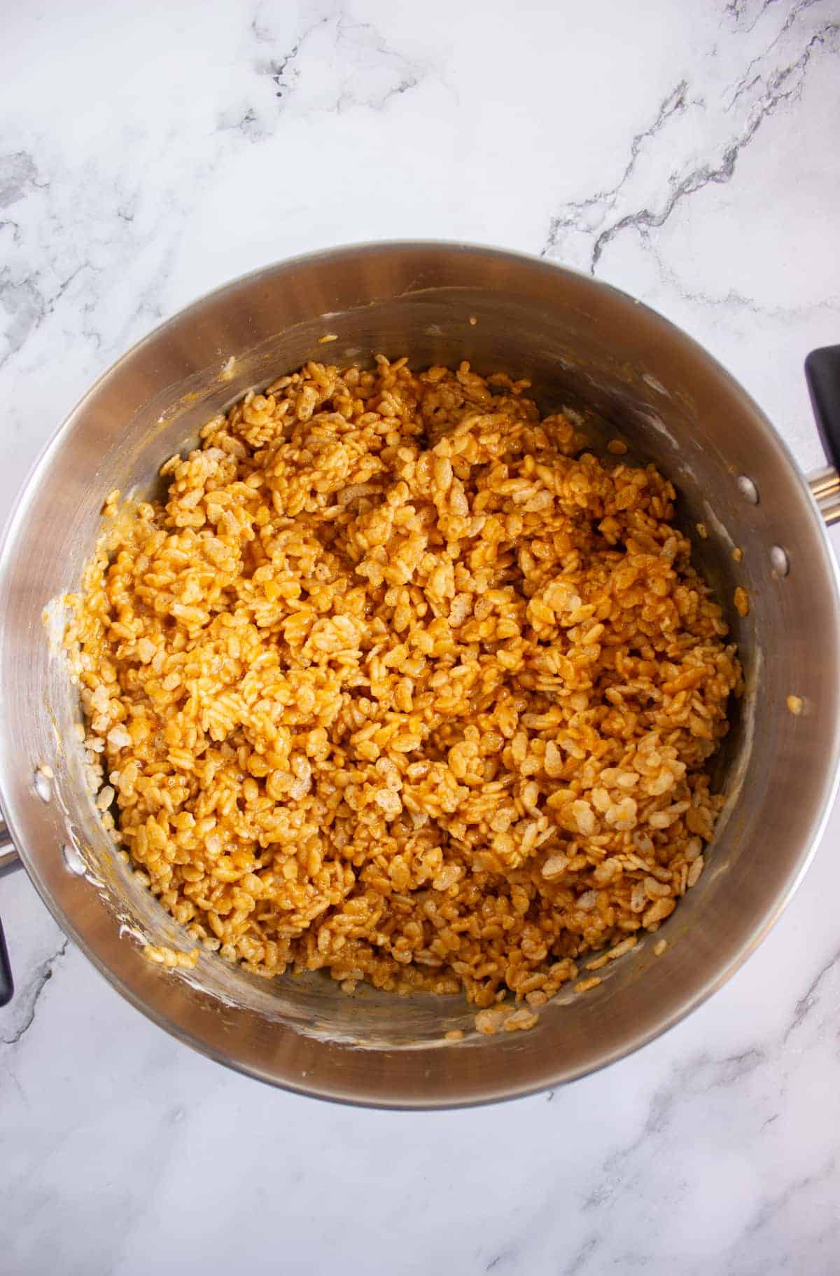 A stainless steel pot filled with a mixture of crispy rice cereal coated in a light brown, sticky substance sits on a marble countertop.