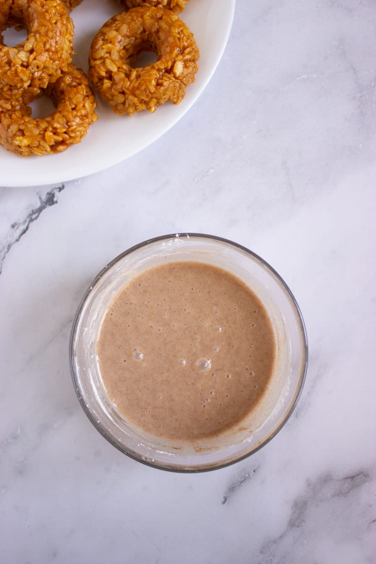 A glass bowl filled with a light brown batter sits on a marble surface, with a plate of round, oat-covered pastries partially visible in the corner.