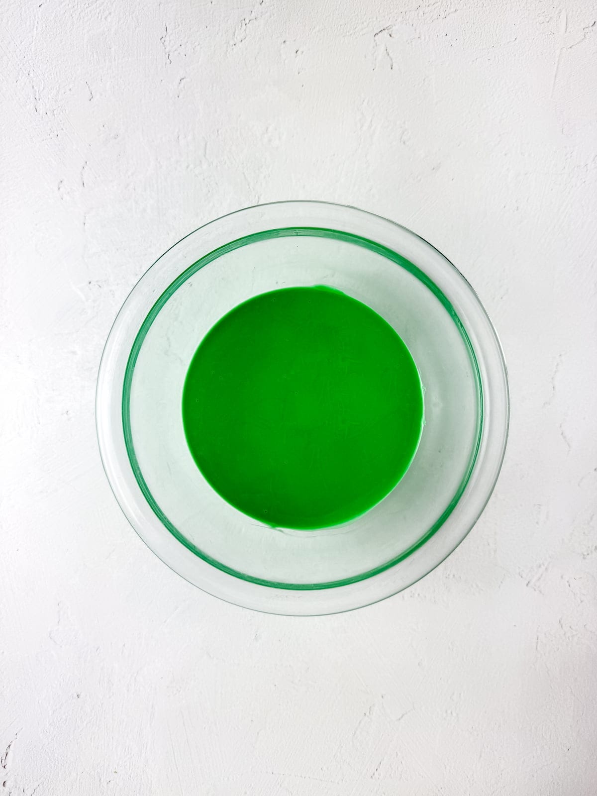 A glass bowl filled with melted bright green chocolate sits on a white textured surface.