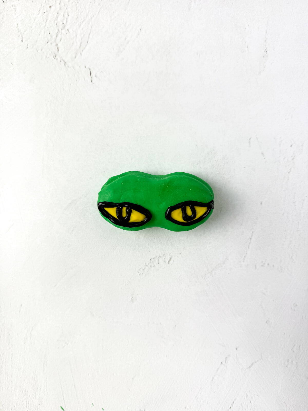 A green object resembling a pair of yellow and black Grinch eyes is placed on a white textured surface.
