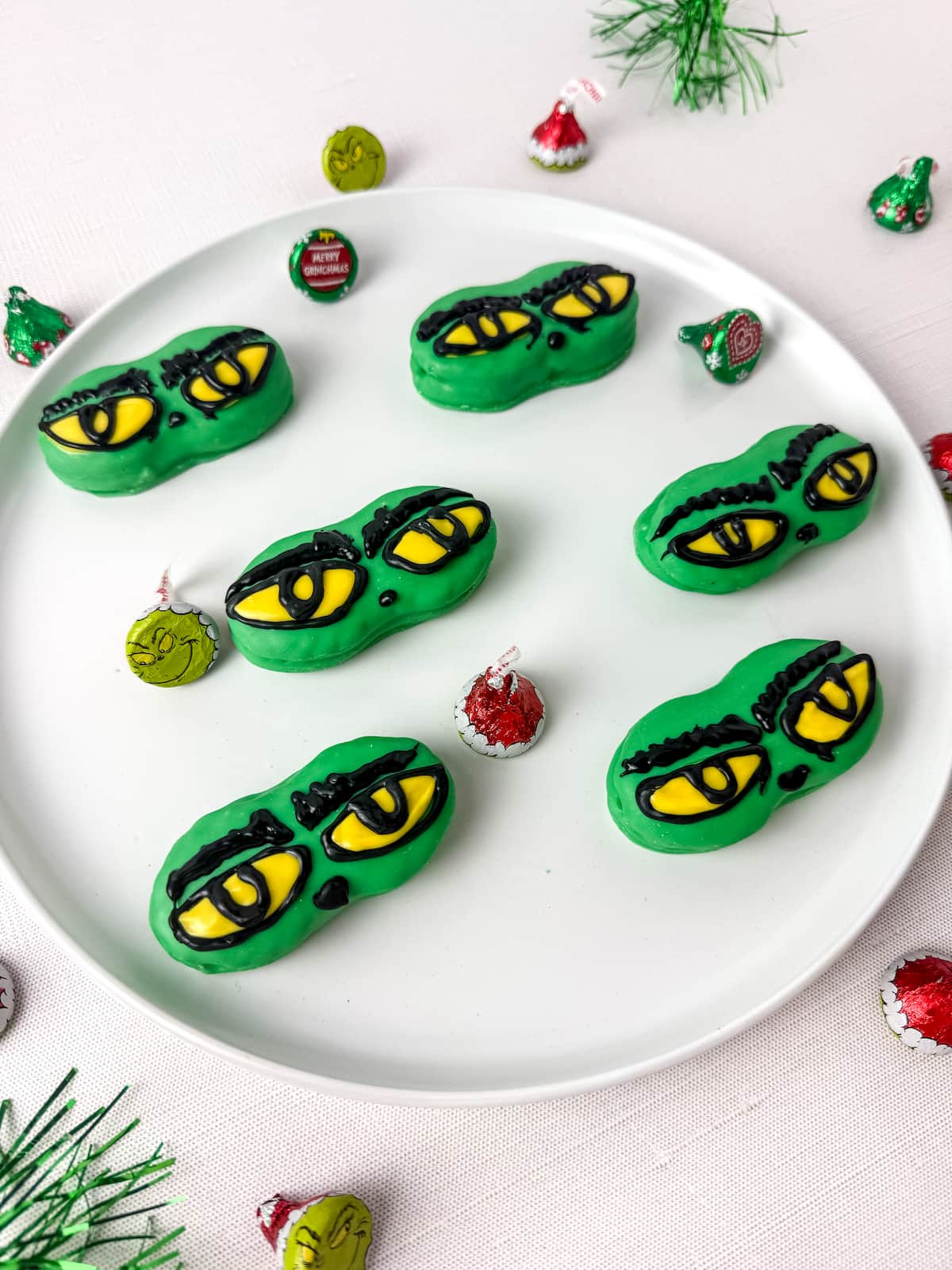 A white plate with six nutter butter cookies decorated with yellow and black eye designs, resembling the Grinch, surrounded by assorted holiday candies.