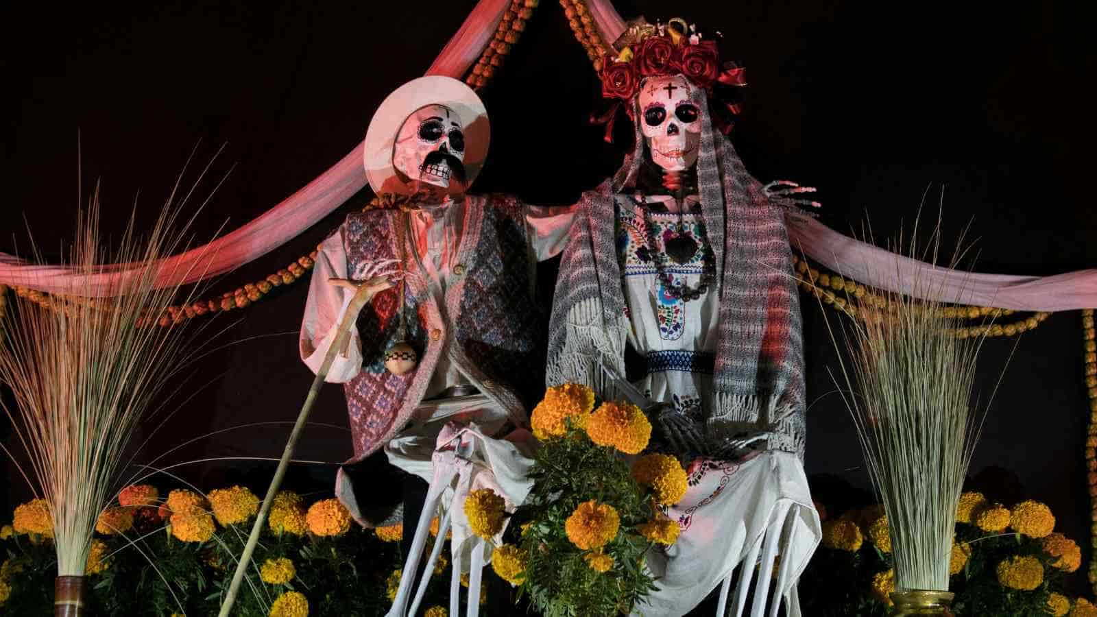 Two life-sized skeleton figures dressed in traditional Mexican attire are displayed with marigold flowers, referencing Día de los Muertos.