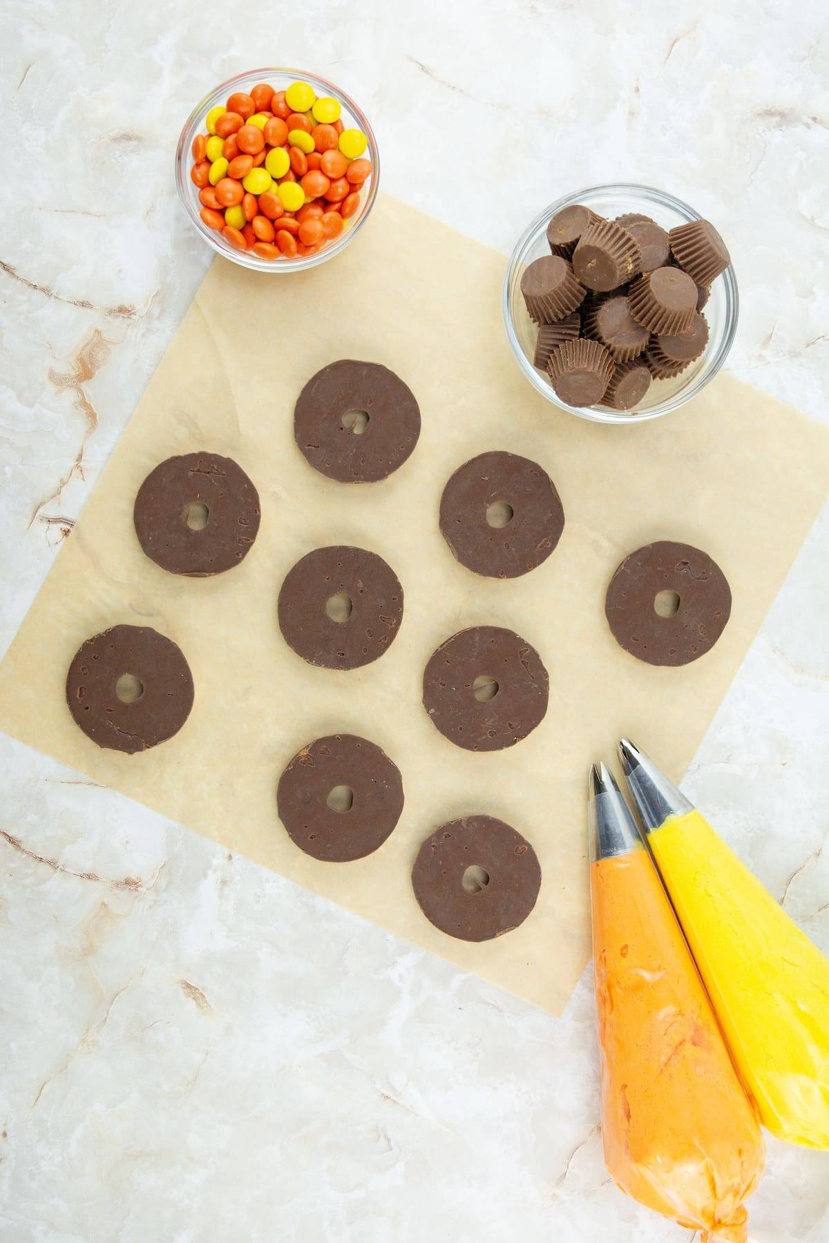 Nine chocolate donut-shaped cookies are arranged on parchment paper, with bowls of mini peanut butter cups and Reese’s Pieces, plus orange and yellow piping bags nearby.