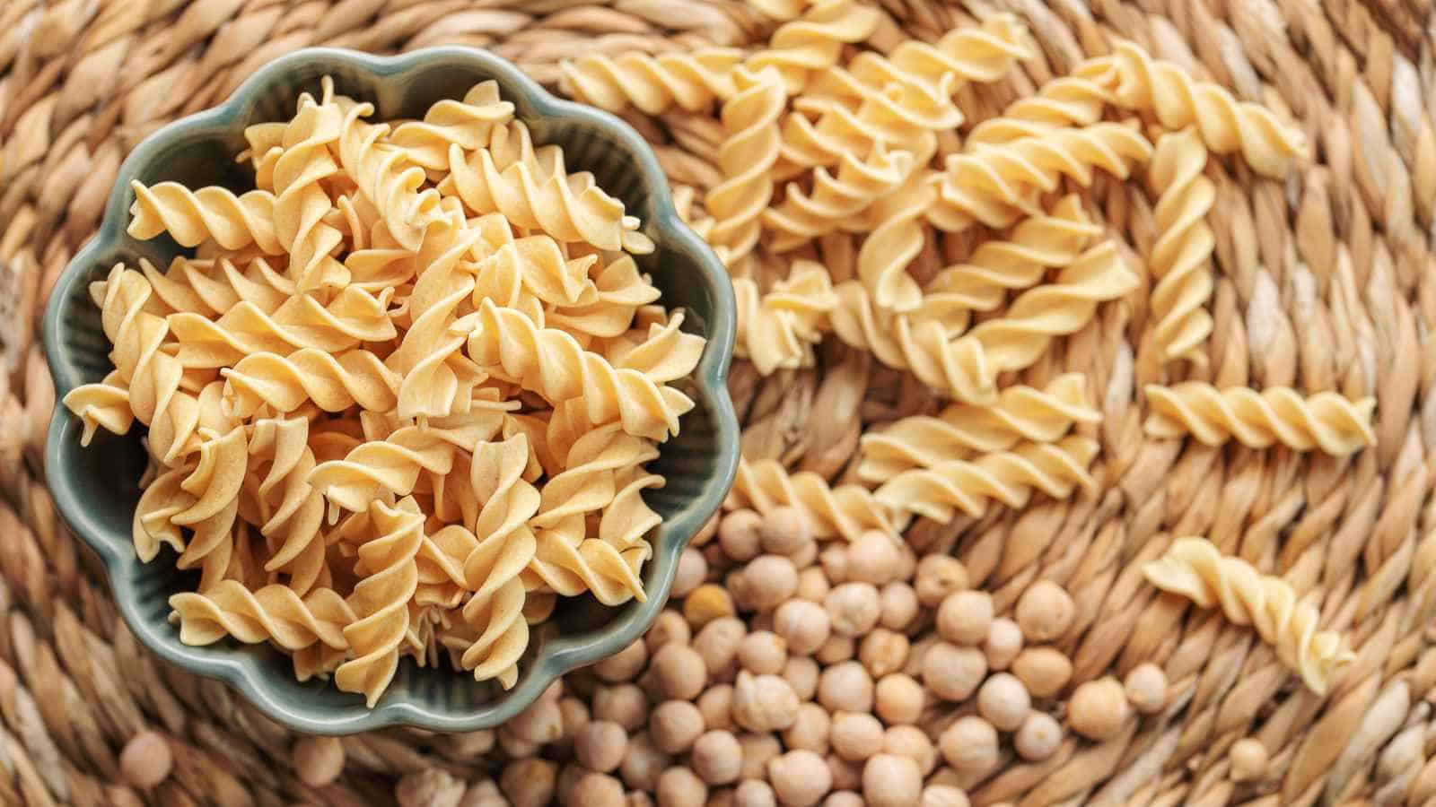 A bowl of uncooked fusilli pasta sits on a woven mat, with some pasta and chickpeas scattered around it.